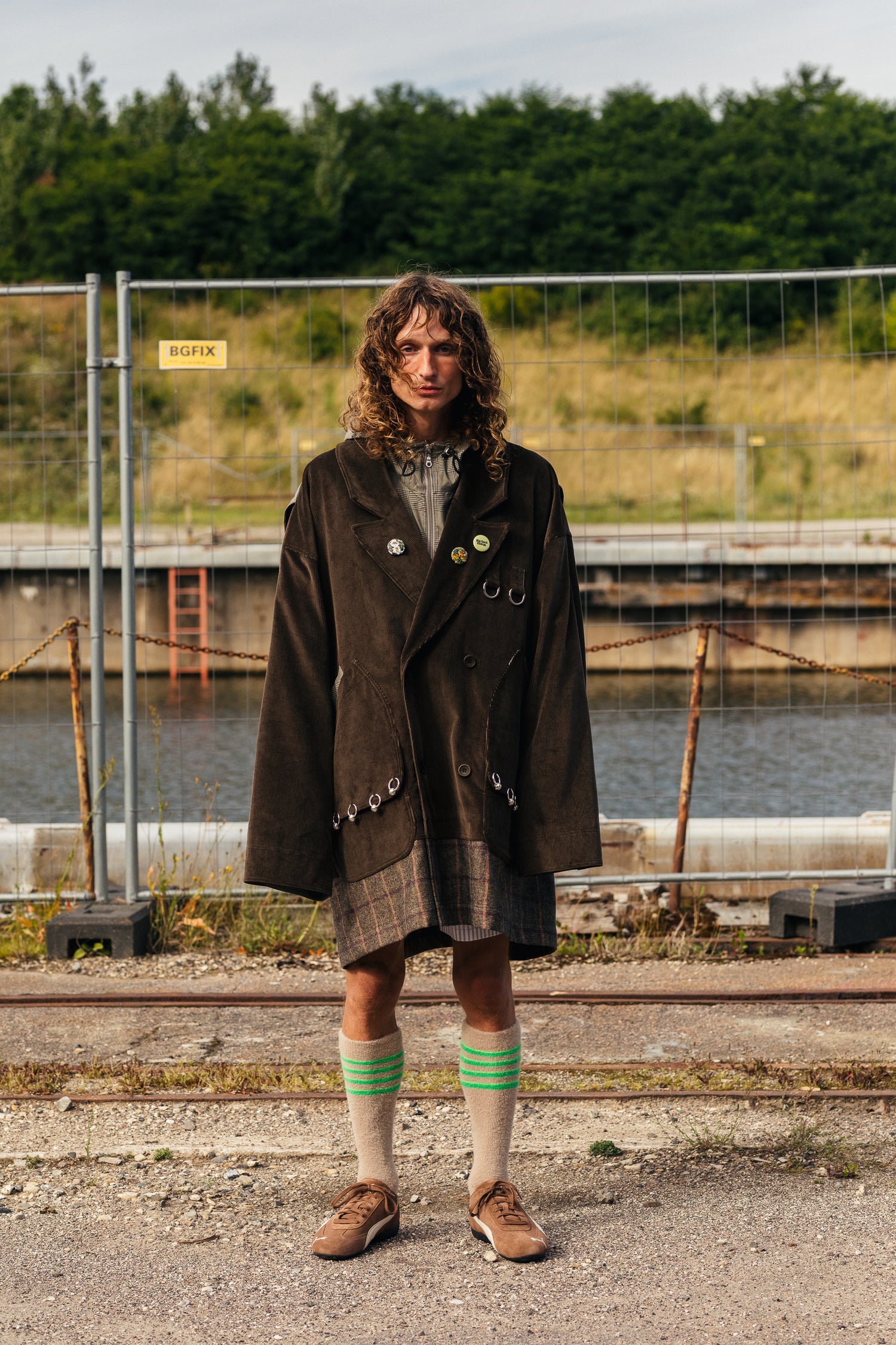 Kiharahiuksisella mallilla on ruskea samettitakki, ruutuhame ja harmaat pitkät villasukat, joissa on vihreitä raitoja. Samettitakin taskuihin on ripustettu kymmenen RAKAS Rolf Ekroth x Kalevala -korua.